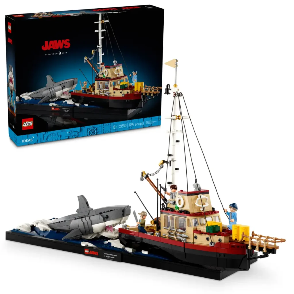 A LEGO set featuring the iconic shark attack scene from the movie "Jaws." The set includes a detailed model of the boat "Orca" with three minifigures representing characters from the film, and a large gray shark lunging out of the water. The box in the background displays the LEGO "Jaws" logo and highlights that the set is part of the LEGO Ideas series, recommended for ages 18+, with a total of 1,497 pieces. The set also features a small plaque with the famous quote, "You're gonna need a bigger boat."