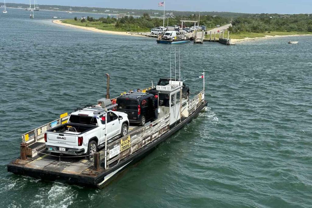 a small car ferry called the On Time III carries 3 vehicles across a small body of water. A ferry dock and sandy beach is visible in the distance. This is one of the fun things to do on chappaquiddick