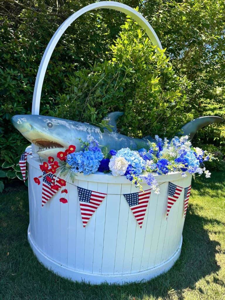 A garden display featuring a large model of a shark emerging from a white planter filled with blue and white flowers. The planter is decorated with small American flag bunting, adding a patriotic theme to the display. The background is lush with greenery.