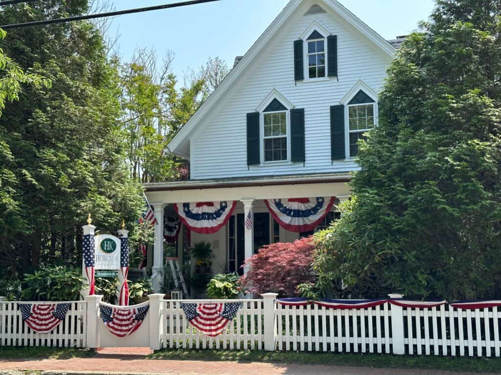 A charming white house, the Hob Knob Inn on Martha's vineyard, adorned with patriotic bunting and American flags on a sunny day, featuring a well-manicured garden and a white picket fence, exuding a quaint, historic ambiance. A good choice for Jaws 50th anniversary on martha's vineyard