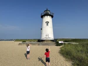 Large white Edgartown Lighthouse against a cloudless sky, viewed up-close with children enjoying the surrounding sandy terrain of Lighthouse Beach