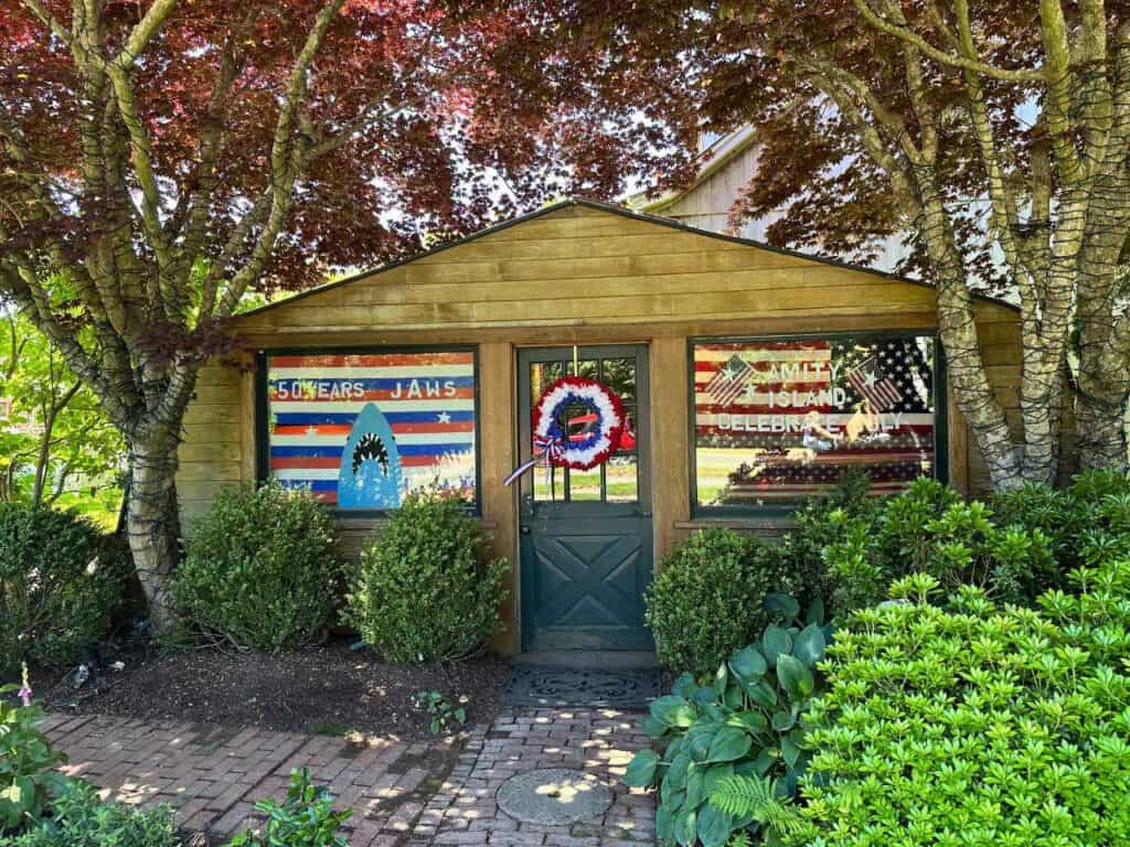 A wooden shed with a window display celebrating the 50th anniversary of the movie "Jaws." The display features a painted shark head on a striped red, white, and blue background. The window includes text that reads "50 Years Jaws" and "Amity Island Celebrate July." The surrounding area is landscaped with trees and bushes.
