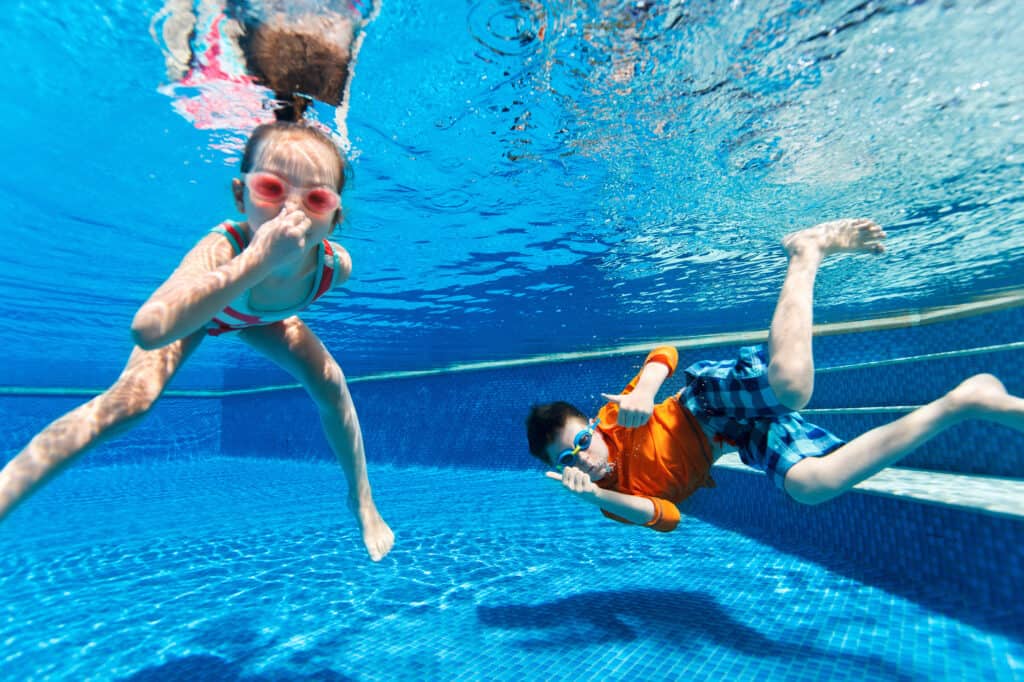 Two children are swimming underwater in a clear blue pool, with one child in striped swimwear and pink goggles leading the way while the other, in orange floaties and blue goggles, follows. Sunlight dances across the surface, casting ripples of light down into the water.