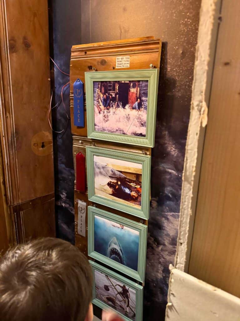 a display of framed photographs on a wooden wall, featuring scenes from the movie "Jaws." The top photo, with a blue "1st place" ribbon, shows people on a boat facing large waves, while the middle images depict the shark attacking and swimming, with a red "2nd place" ribbon and a grey "3rd place" ribbon beside them. The display has a rustic and nautical theme