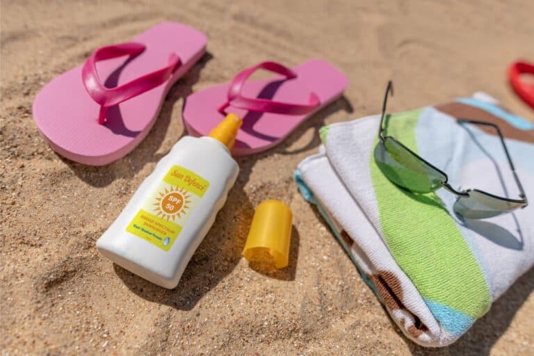 a bottle of sunscreen, pink flip flops and a folded beach towel with sunglasses on top, rest on the sand