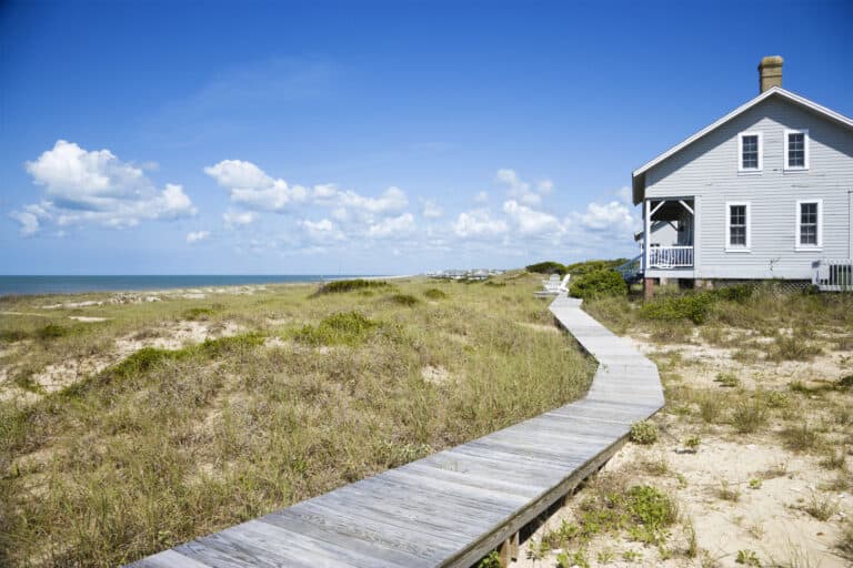 A serene beachside getaway featuring a weathered wooden boardwalk leading to a charming white coastal cottage like a chappaquiddick vacation rental with a wraparound porch, nestled among wild grasses under a clear blue sky with fluffy clouds