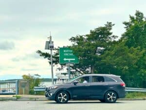A dark gray SUV waits at an intersection with a green road sign pointing left toward “Martha's Vineyard”. In the background, a ferry and the waterfront are visible beneath a cloudy sky, with lush green trees lining the roadside. A stone marker engraved with “1952” stands near the edge of the road.
