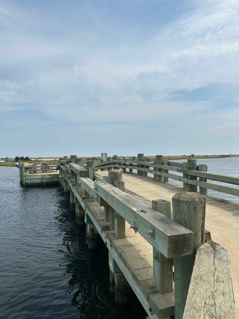 a wooden bridge with a sandy surface and thick wooden rails. water can be seen underneath and on either side