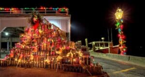 Christmas-decorated lobster trap tree at a New England harbor dock, made from stacked copper-colored traps and adorned with colorful lights and buoys. A red fishing boat is visible in background, while a decorated lamppost with multicolored lights and evergreen garland stands at right, creating festive maritime scene at night. Martha's Vineyard in December