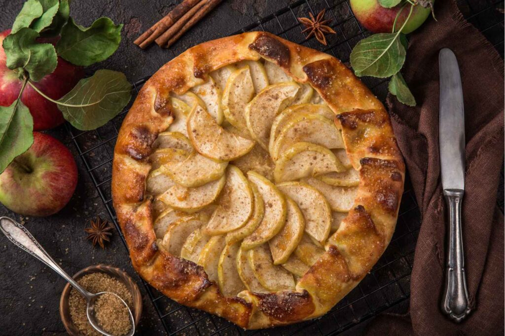 an overhead picture of an apple pie. Appleas are arranged in slices on top of the pie with pastry folding inwards at the edges. A knofe is on the right, some apples with stems and leaves on the left and cinnamon sticks, a spoon in some ground spices and whole star anise scattered around the pie