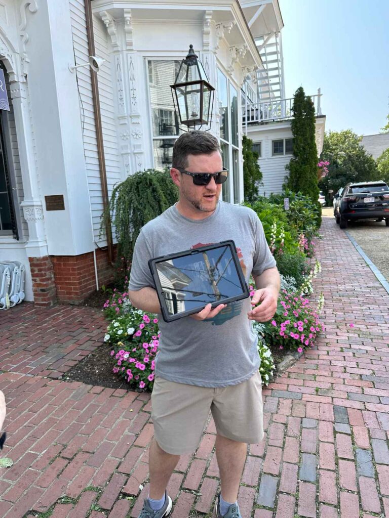 a man wearing sunglasses, shorts and grey tshirt holds an ipad. It is facing away from him. He stands on a redbrick pavement with a white house in the background. he is a tour guide on a Martha's Vineyard Jaws tour