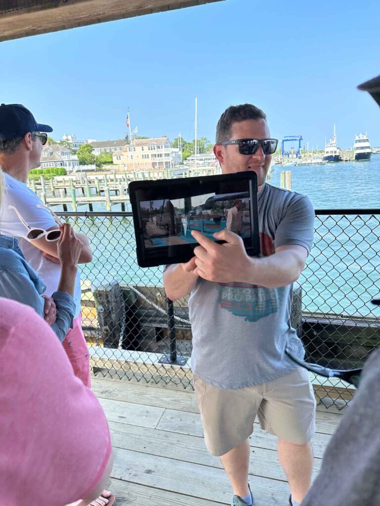a man holds an ipad with the screen facing away from him. He is pointing at an image on the screen. It is a still from the movie Jaws. he is a tour guide on one of Martha's Vineyard jaws tours. Edgartown Harbor is in the background