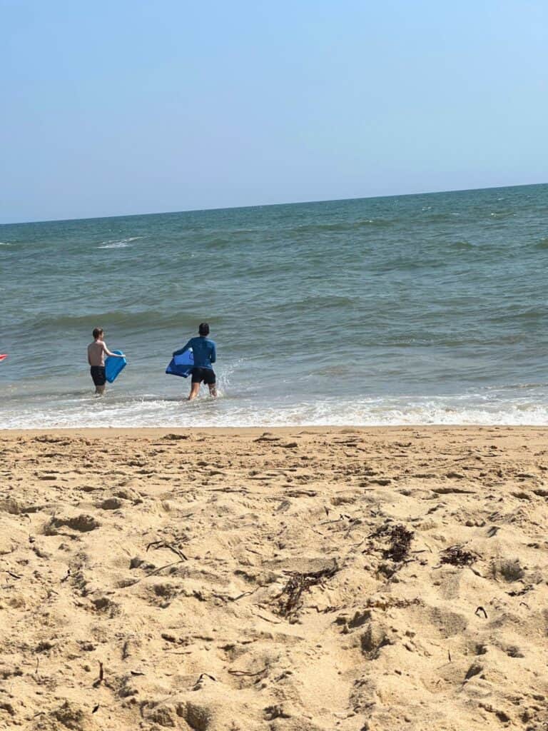 Two people, one in a blue wetsuit, playfully engage with the rolling waves of the ocean, with boogie boards in hand, on the sandy beachfront of South Beach