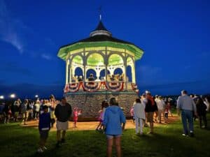 A lit gazebo in Oak Bluffs, adorned with patriotic bunting, is the focal point of a nighttime gathering. People stand around the gazebo, which glows warmly against the deep blue evening sky