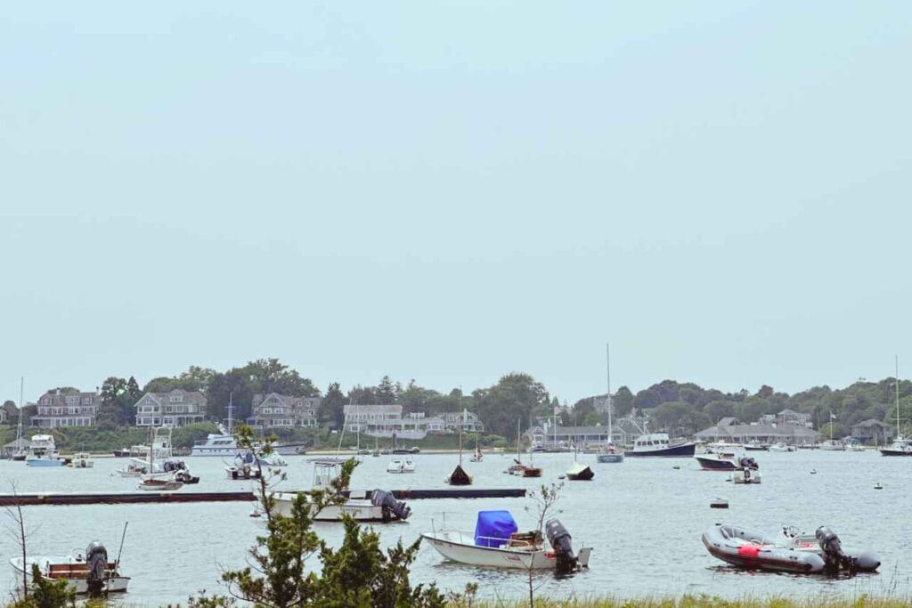 A serene view of edgartown from Chappaquiddick with a variety of boats moored on calm waters. In the background, grand houses overlook the scene, nestled among lush greenery, under a hazy sky